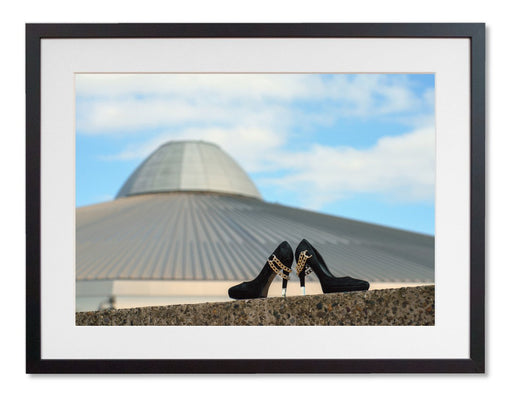 A framed print with a black frame, the print showing a pair of high heel shoes on a wall in front of a building