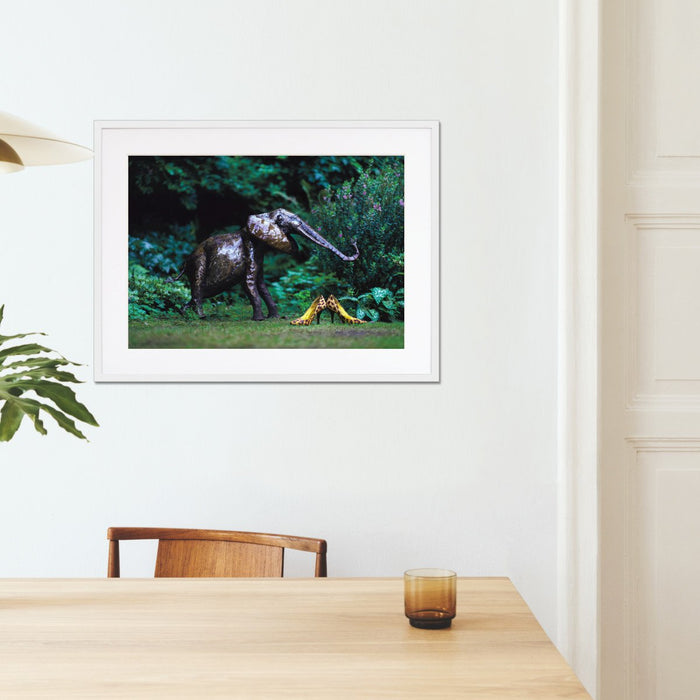 A framed print with a white frame hung on the wall of a kitchen, the print showing pair of yellow high heel shoes in a garden next to a garden elephant