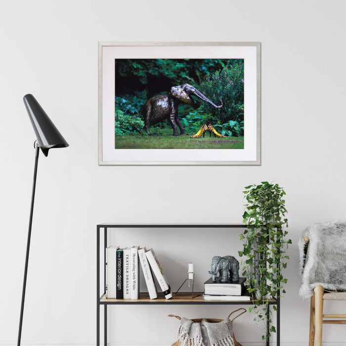 A framed print with a silver frame hung on the wall of a living room, the print showing pair of yellow high heel shoes in a garden next to a garden elephant