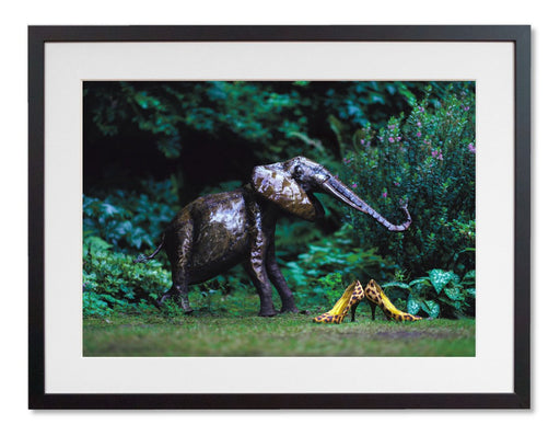 A framed print with a black frame, the print showing pair of yellow high heel shoes in a garden next to a garden elephant