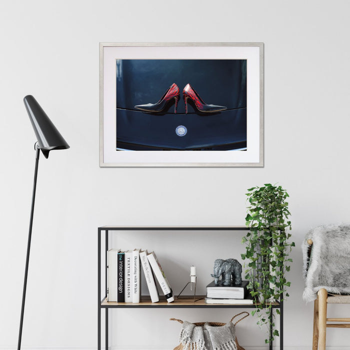 A framed print with a silver frame on the wall of a living room, the print showing a pair of purple high heel shoes on the bonet of a high performance car