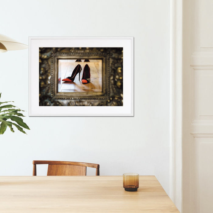 A framed print with a white frame hung on the wall of a kitchen, the print showing a pair of orange and black high heel shoes reflected in a mirror