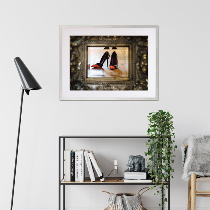 A framed print with a silver frame hung on the wall of a living room, the print showing a pair of orange and black high heel shoes reflected in a mirror