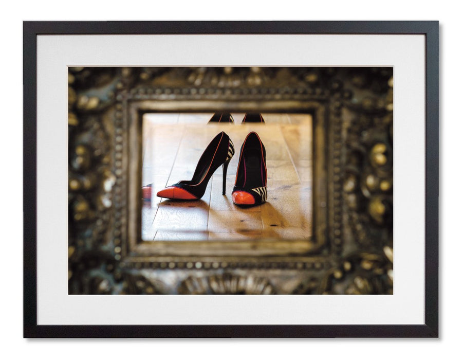 A framed print with a black frame, the print showing a pair of orange and black high heel shoes reflected in a mirror