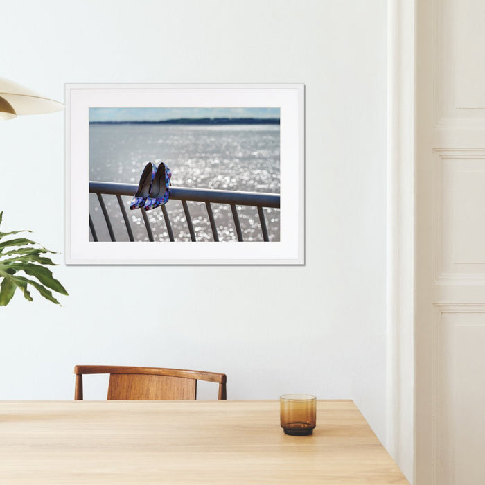 A framed print with a white frame hung of the wall of a kitchen, the print showing a pair of blue high heel shoes hung off a metal fence, in front of the ocean