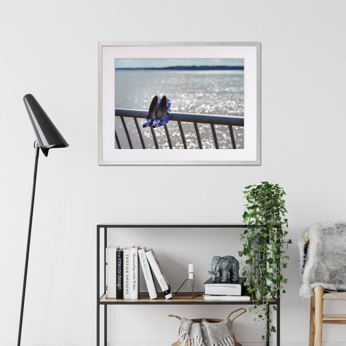 A framed print with a silver frame hung on the wall of a living room, the print showing a pair of blue high heel shoes hung off a metal fence, in front of the ocean