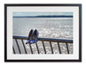 A framed print with a black frame, the print showing a pair of blue high heel shoes hung off a metal fence, in front of the ocean