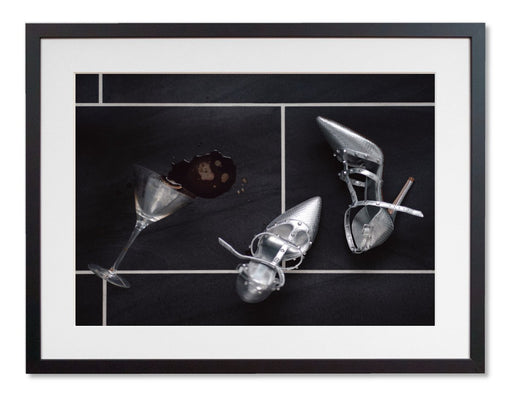 A framed print with a black frame, the print showing a pair of silver high heel shoes lying on a tiled floor next to a spilt cocktail drink