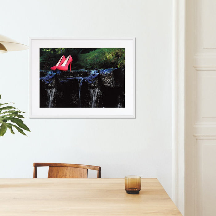 A framed print with a white frame hung on the walls of a kitchen, the print showing a pair of pink high heel shoes sat on a rock in a flowing river