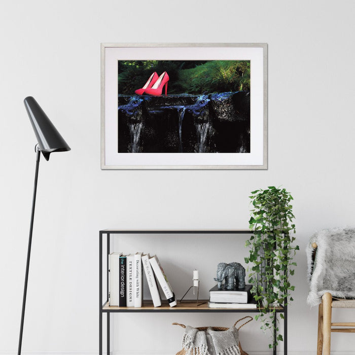 A framed print with a silver frame hung on the wall of a living room, the print showing a pair of pink high heel shoes sat on a rock in a flowing river