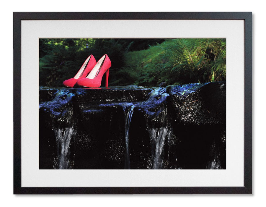A framed print with a black frame, the print showing a pair of pink high heel shoes sat on a rock in a flowing river