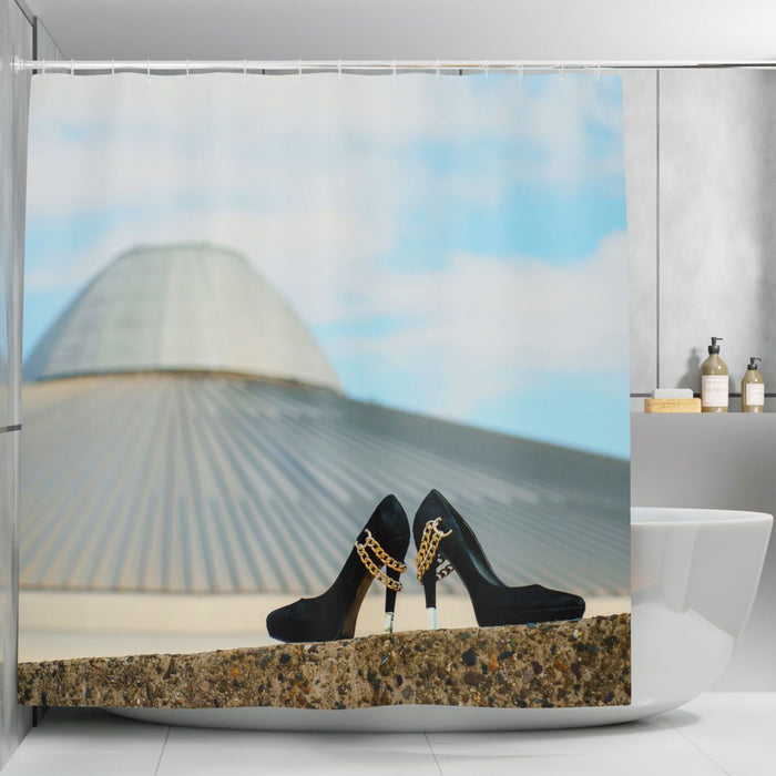 A shower curtain in a bathroom, the shower curtain having an image of a pair of black high heel shoes on a wall in the foreground, with a building and city skyline in the background