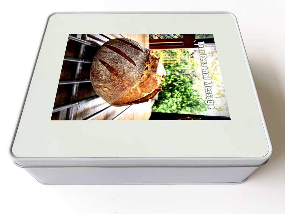 A white tin box, on the lid of the box is an image showing an image of a large sourdough loaf on  a table with a garden visible behind through the window