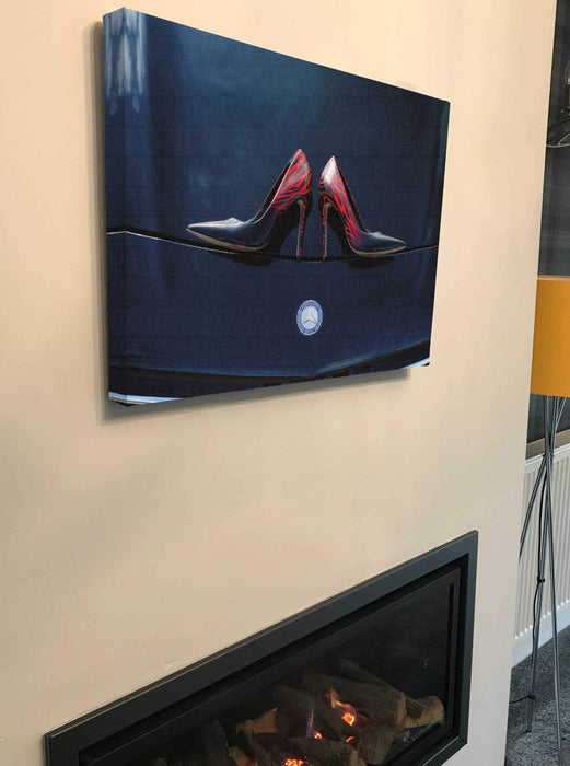 A canvas print of a pair of blue high heel shoes sitting on the bonet of a high powered card, the canvas print is hung above a fire place and seen from a side angle