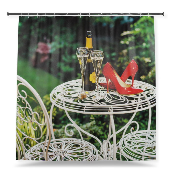 A shower curtain showing an image of a pair of red high heel shoes sat on a white garden table next to a bottle of fizzy wine with two poured glasses of fizz, with a white background