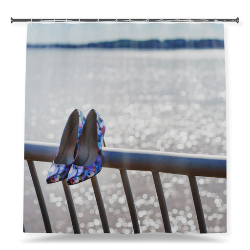 A shower curtain showing an image of a pair of blue high heel shoes hooked on the top of some metail railings in the front of the ocean, with a white background