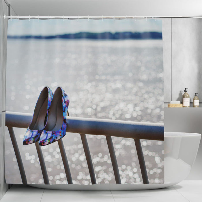 A shower curtain in a bathroom, the shower curtain having an image of a pair of purple high heel shoes hung on metail railings in front of the ocean