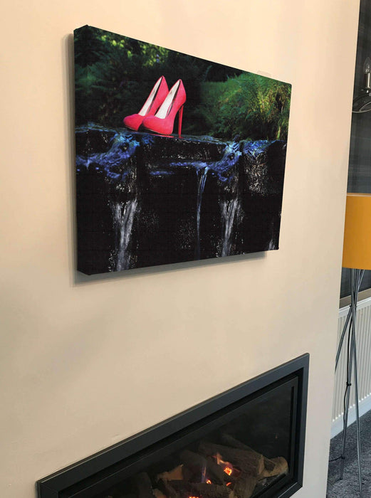 a canvas print of a pair of pink high heel shoes hald way across a river and resting on a rock, the canvas hung above a lit fire in a living room