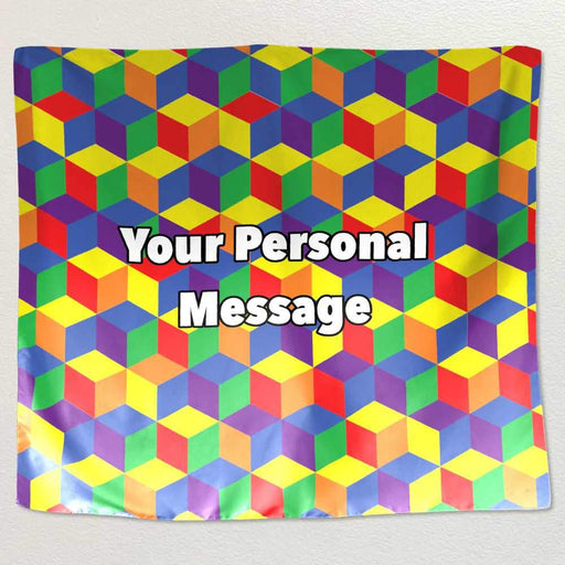 A tapestry hung on a white wall, the tapestry has 3D cubes on it, each cube face coloured with a rainbow colour, along with a printed personal message
