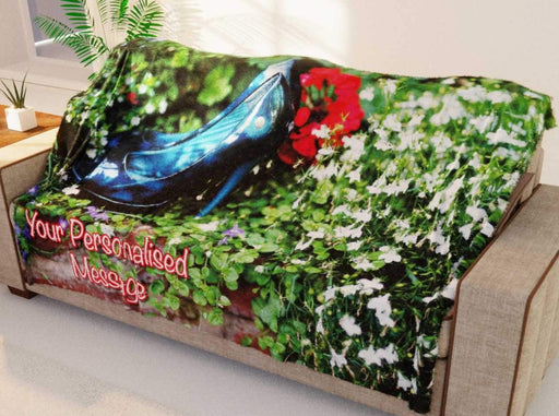 A blanket showing an image of a pair of blue high heel shes in a flower bed full of flowers, the blanket is draped over a couch in a living room