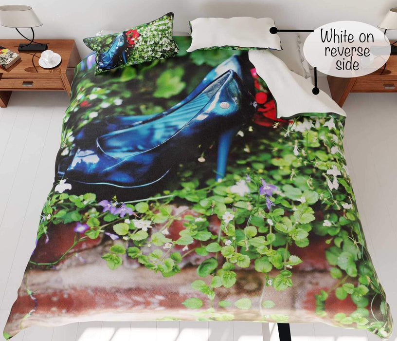 A bed with a duvet cover, the duvet having an image of a pair of blue high heel shoes surrounded by flowers, the corner of the duvet is folded over to show a white underside