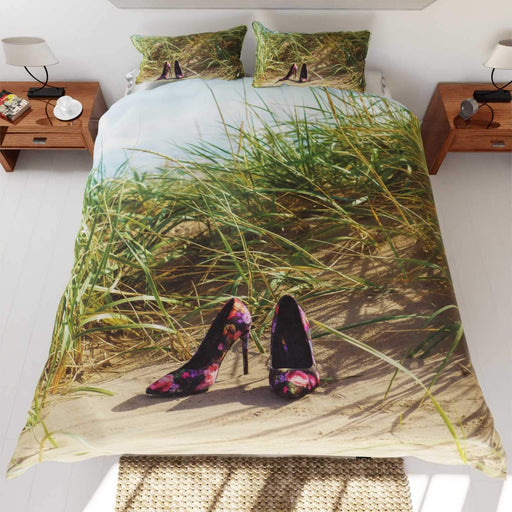 A duvet cover on a bed, the duvet cover having an image of a pair of high heel shoes on a beach near to some sandhills with grass and blue sky