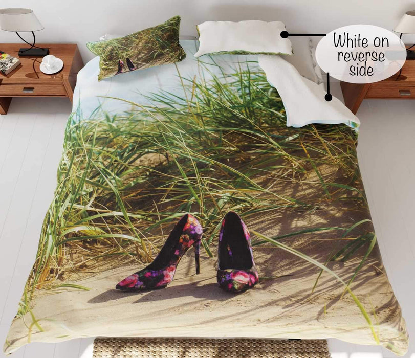 A duvet cover on a bed, the duvet cover having an image of a pair of high heel shoes on a beach near to some sandhills with grass and blue sky, with a printed personal message on the duvet