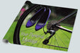A roll of wrapping paper showing the rear wheel of a racing bicycle with a pair of purple heels hung from the spokes of the wheel, there is grass and trees in the background, and a personal message