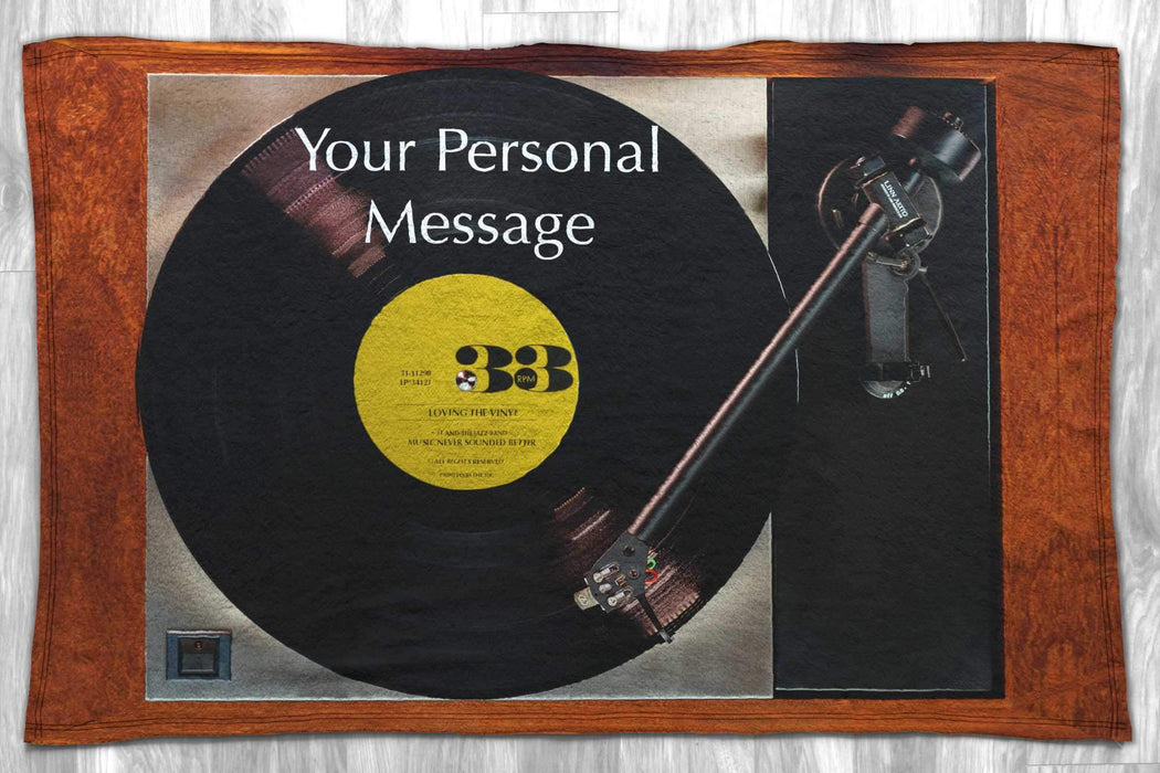 A blanket laid down on a wooden floor, the blanket showing an image for a record player with a vinyl record playing with a yellow label