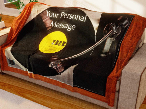 A blanket draped over a couch, the blanket having an image of a record player with a yellow label vinyl record playing on it
