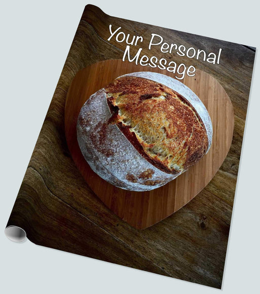 A roll of wrapping paper showing a sourdough bread loaf sat on a woodern tray in the shape of a heart, upon a brown table, with a personal message