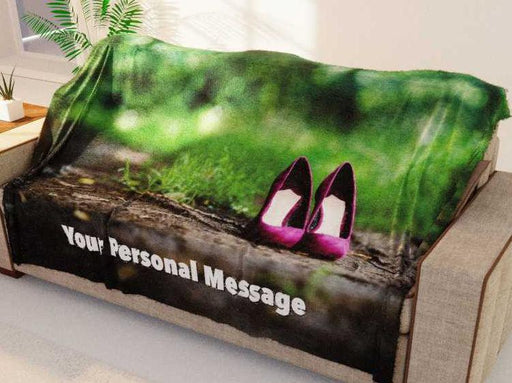 A blanket with an image of a pair of purple high heel shoes standing on a woodland path, the blanket is draped over a couch and there is a personal message printed