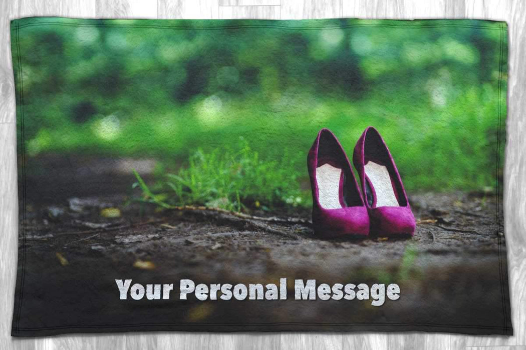 A blanket with an image of a pair of purple high heel shoes standing on a woodland path, the blanket is flat on the floor and there is a personal message printed