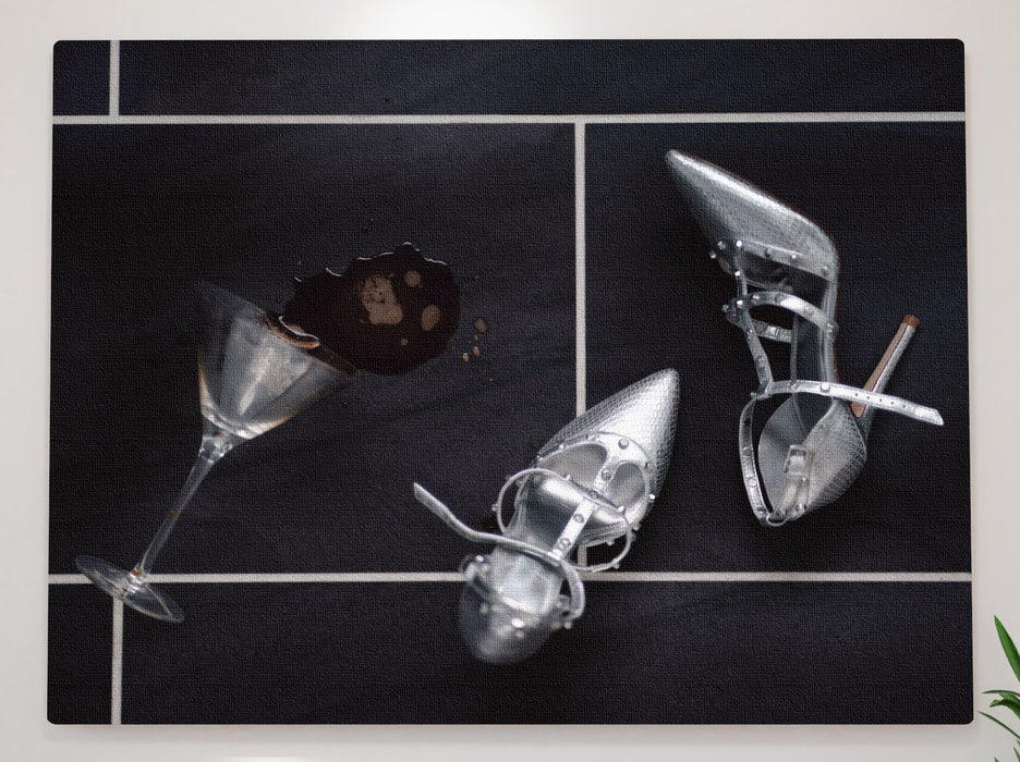 An image of a spilt cocktail drink with a fallen cocktail glass on a black floor, with a pair of silver high heel stilettos lying next to the spilt drink
