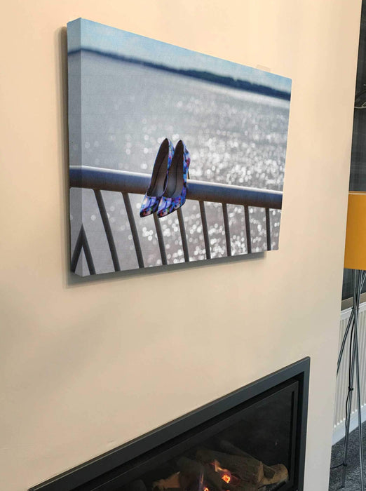 A canvas print of a pair of purple shoes sat on a blue metal fence on the shore of an ocean, with the ocean sparkling in the background, the canvas print is hung above a fire place in a living room
