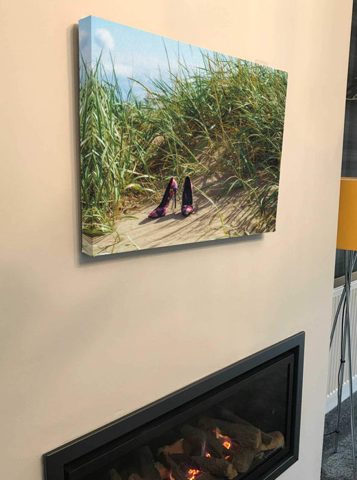 A canvas print of a pair of multicoloured shoes sat on sand on a beach, with sand dunes with grass in the background, the canvas print is hung above a fire place in a living room and seen from a side angle