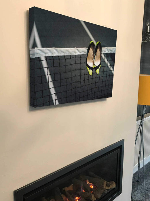 A canvas print of a pair of yellow high heel stiletto shoes hung on a tennis net, the canvas print is hung above a fire place