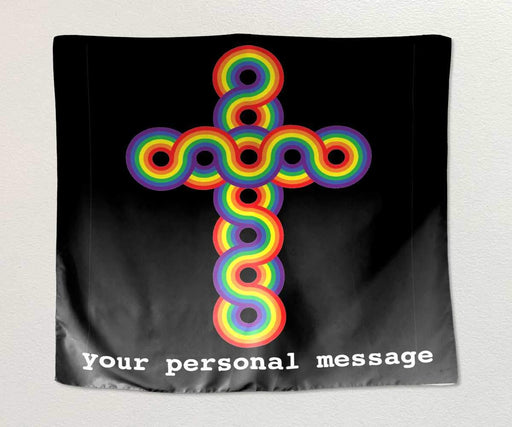 A tapestry hung on a white wall, the tapestry has a rainbow coloured crucifix on a black background, along with a personal message printed