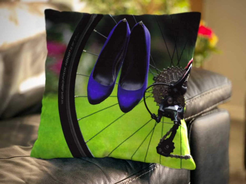 An image of a cushion on a couch, the cushion cover showing an image of a rear wheel of a bicycle with a pair of purple high heel shoes hung over the spokes of the wheel