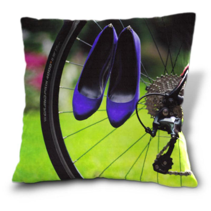An image of a cushion, the cushion cover showing an image of a rear wheel of a bicycle with a pair of purple high heel shoes hung over the spokes of the wheel