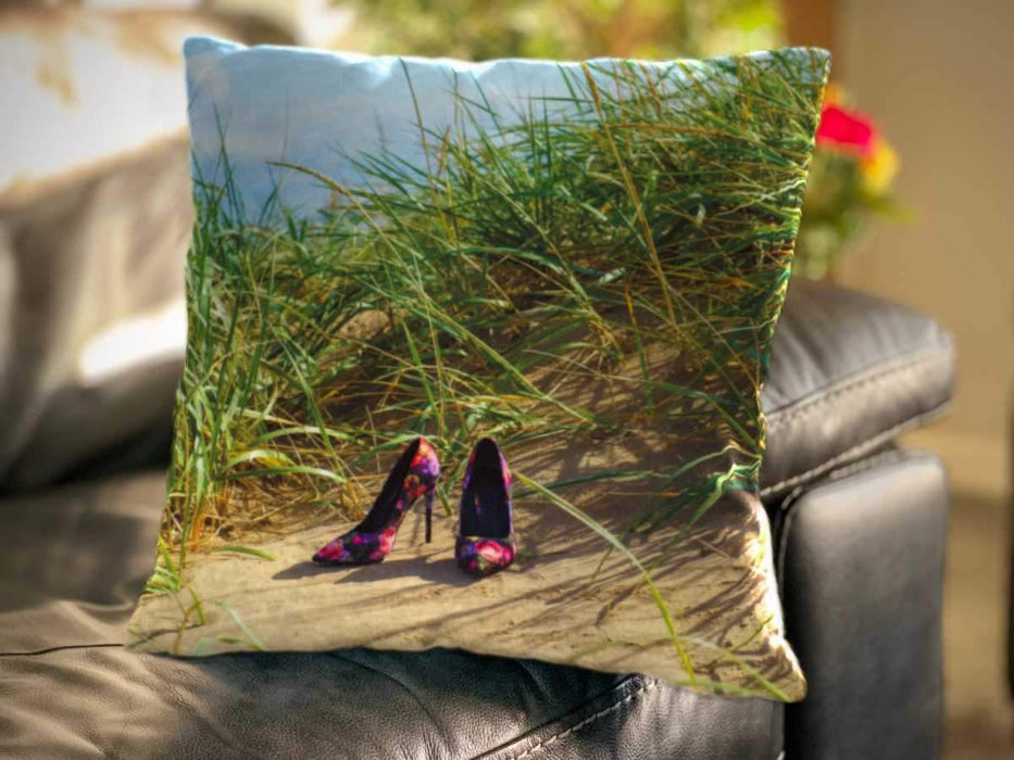 An image of a cushion sat on a couch, the cushion having an image of a pair of multicoloured high heel shoes sat on a beach in front of a grassy sand hill