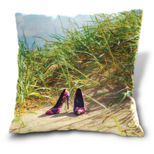 An image of a cushion, the cushion having an image of a pair of multicoloured high heel shoes sat on a beach in front of a grassy sand hill