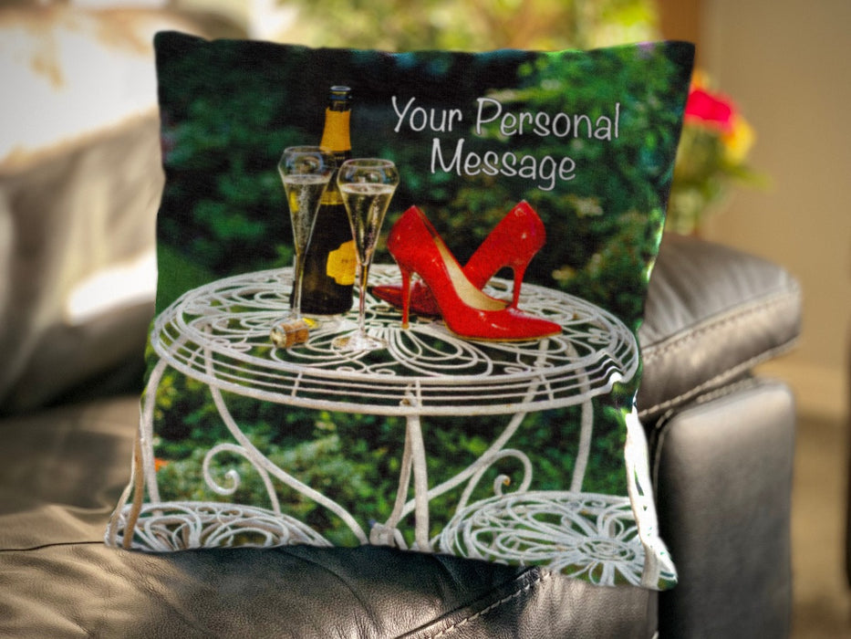 A cushion on a couch, the cushion having an image of a pair of red shoes on a white metal garden table, sat next to a bottle of fizzy wine and two poured glasses, along with a perosnal message on the cushion