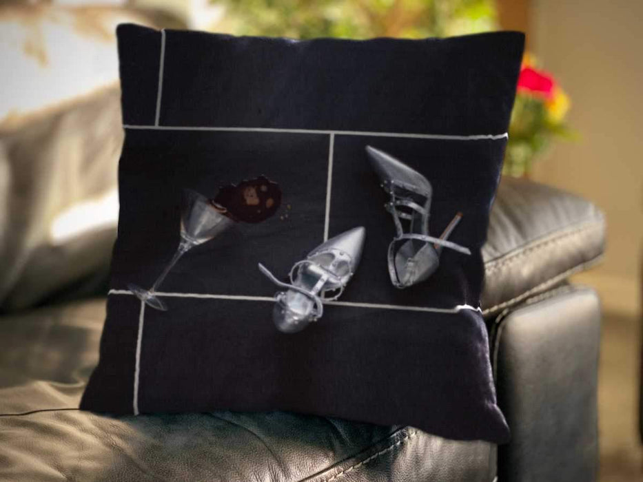 An image of a cushion sat on a couch, the cushion cover showing a cocktail glass on its side with a spilt drink on the floor next to a pair of silver high heel shoes