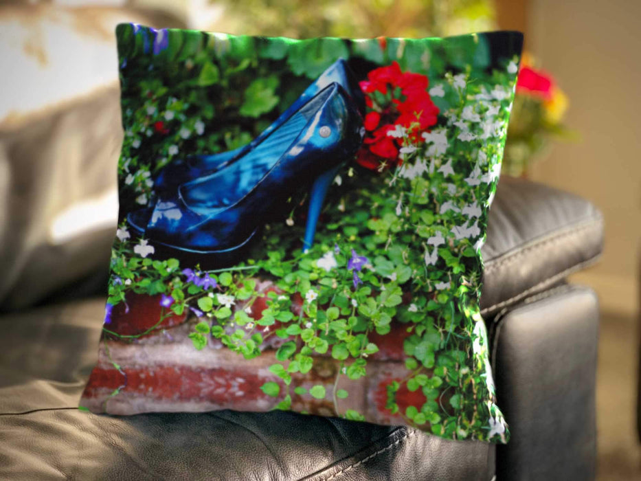 An image of a cushion on a couch, the cushion cover displaying a pair of blue high heel shoes sat inside a bunch of flowers and leaves