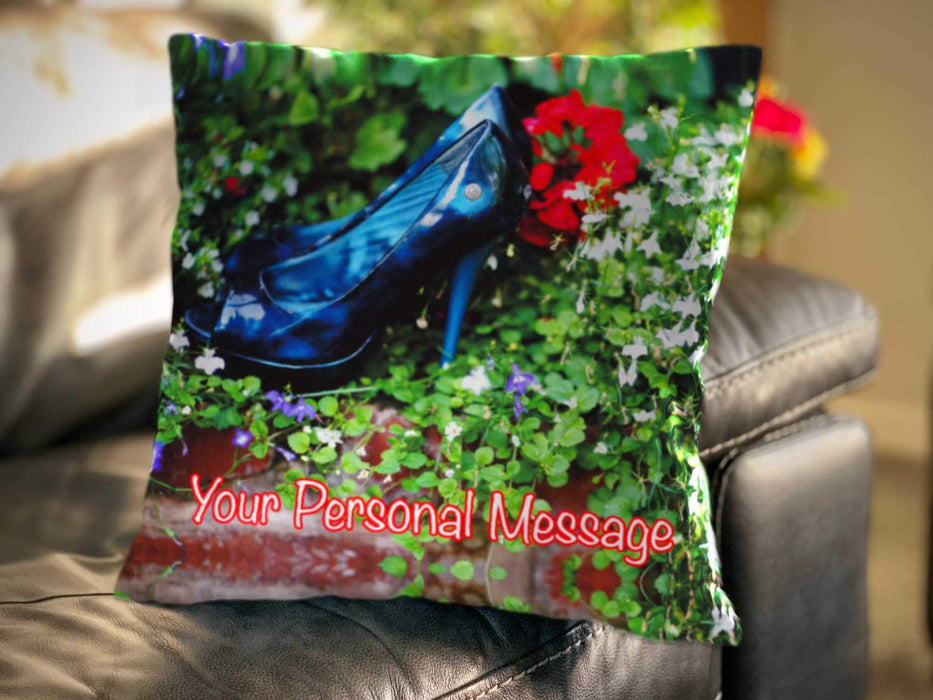 An image of a cushion on a couch, the cushion cover displaying a pair of blue high heel shoes sat inside a bunch of flowers and leaves, along with a personal message on the cushion