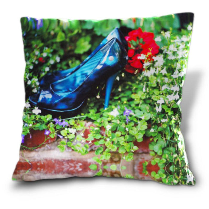 An image of a cushion, the cushion cover displaying a pair of blue high heel shoes sat inside a bunch of flowers and leaves