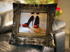 An image of a cushion sat on a couch, the cushion cover showing a mirror with a pair of orage and black high heel shoes reflected in it