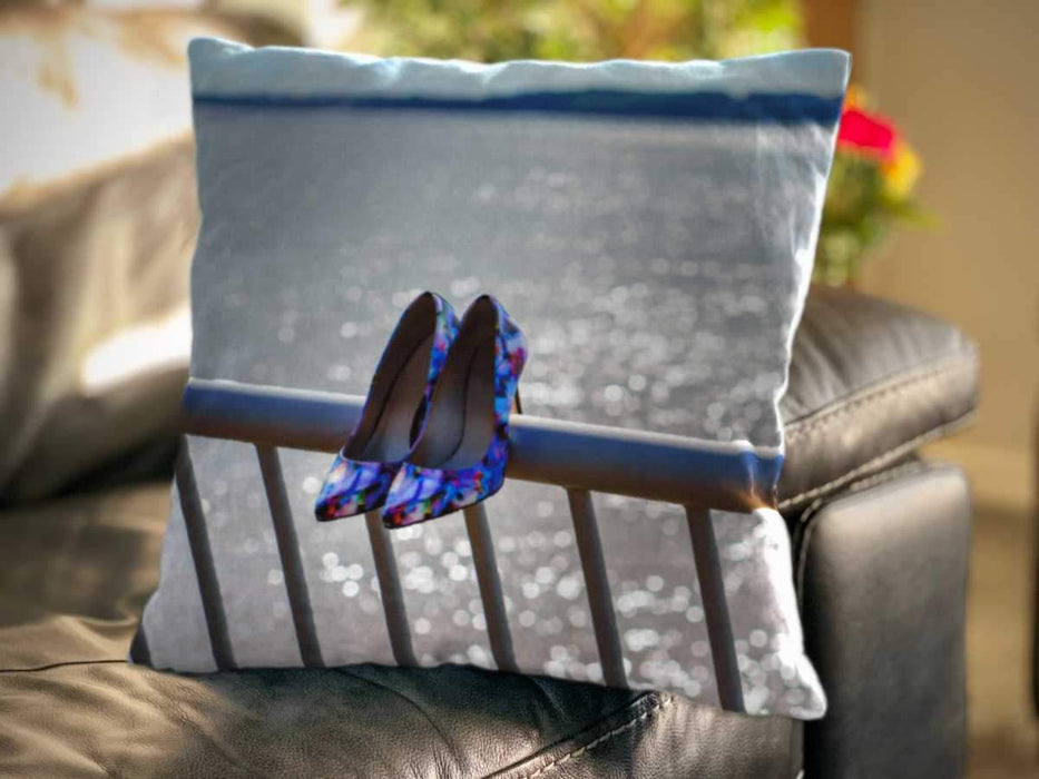 An image of a cushion on a couch, the cushion showing an image of a pair of blue shows hung on the railings adjacent to ocean with the water sparkling behind