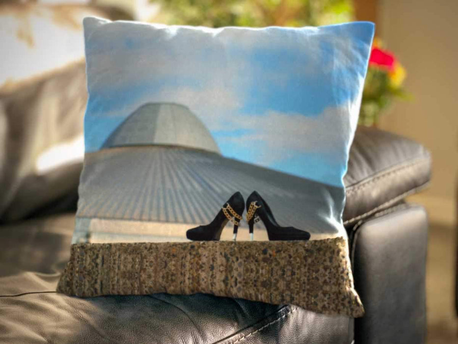 An image of a cushion, the cushion sat on a couch and having a cover showing a pair of black high heel shoes sat on a wall in front of a building with blue sky behind it